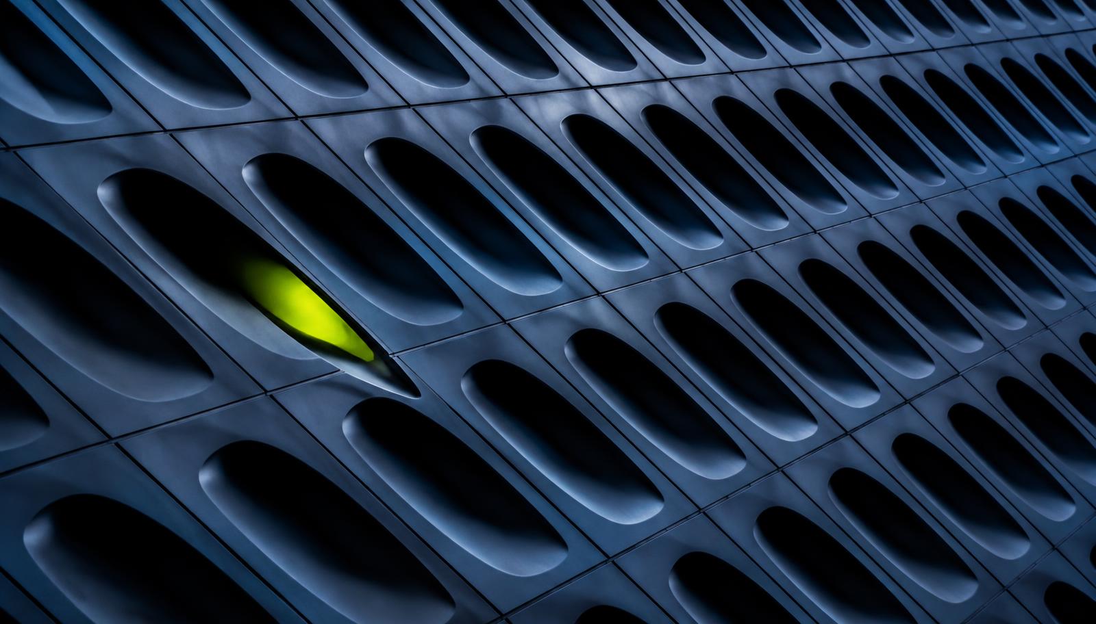 Abstract close-up of a dark blue engineered panel with repeating apertures and a single lime-lit opening